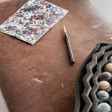A close-up of an artist's workspace featuring a brown leather pad, a paint palette with mixed colors, a fine detail brush, and a foam tray holding small round paint containers.