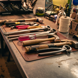 A collection of hand tools, including hammers, pliers, and screwdrivers, laid out on a brown leather work mat on a metal workbench.
