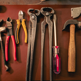 A set of hand tools arranged on a dark brown leather surface, including metal angle clamps, pliers with red and yellow handles, a screwdriver with a pink and purple handle, and a hammer with a wooden handle.