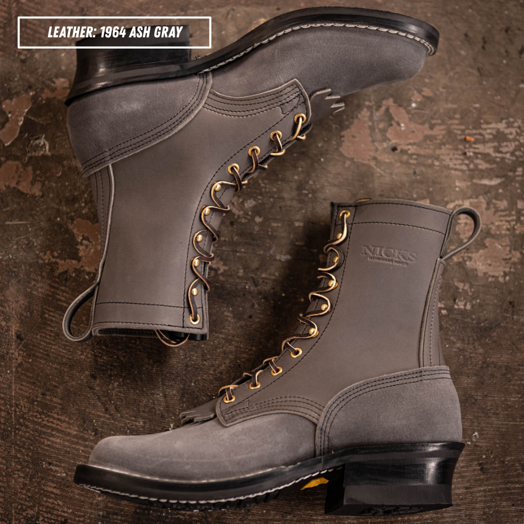 Two gray leather lace-up boots arranged on a rustic brown textured surface. One boot is positioned upright and the other lies on its side, showing the side profile and toe.