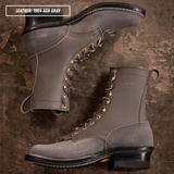 Two gray leather lace-up boots arranged on a rustic brown textured surface. One boot is positioned upright and the other lies on its side, showing the side profile and toe.