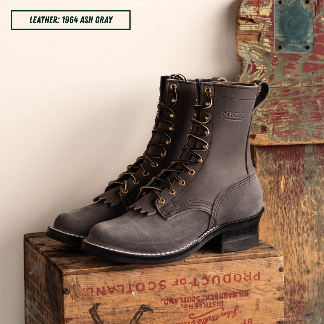 A pair of dark gray leather lace-up boots with fringed detailing and brass eyelets, displayed on a rustic wooden crate against a cream wall and weathered wood panel.