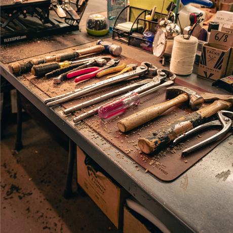 A workbench scene with multiple hand tools laid out on a brown leather mat, including hammers, pliers, screwdrivers, and a metal wrench. The bench is cluttered with workshop items, and the scene is lit with warm, somewhat dim lighting.