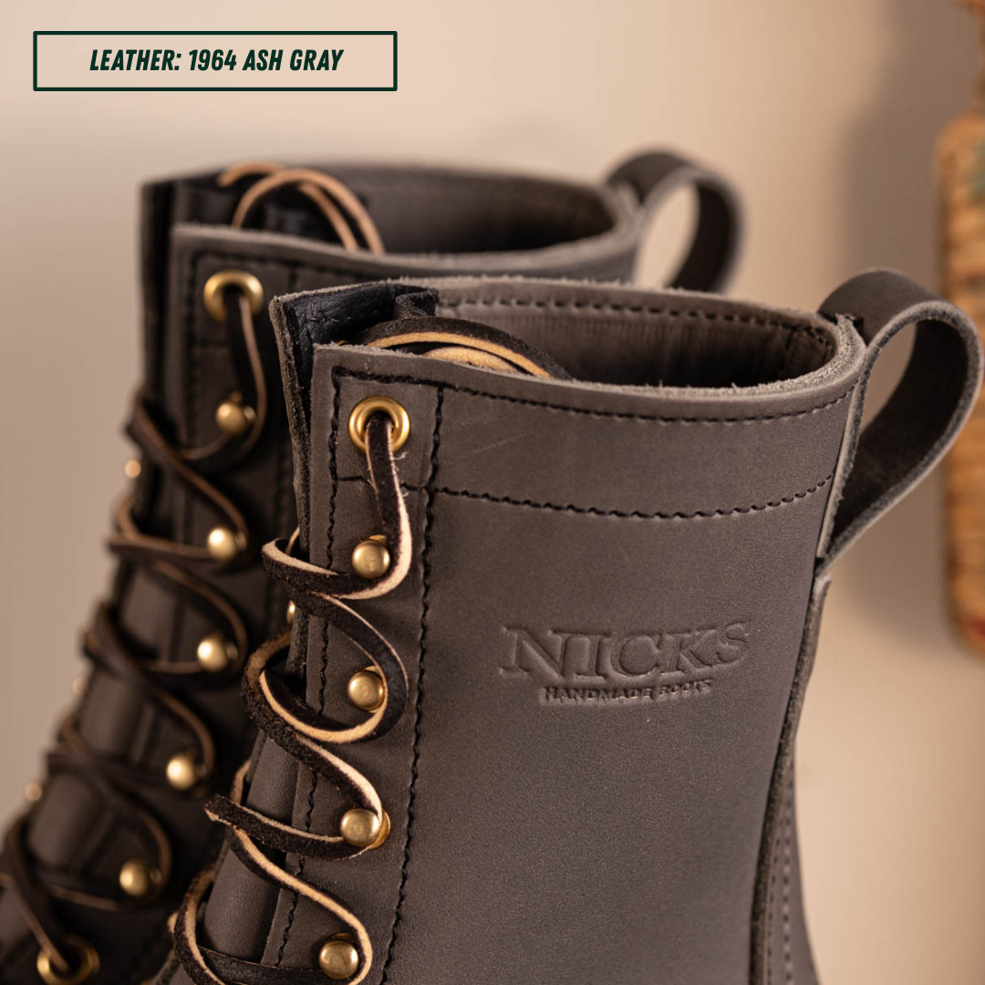 Close-up of a pair of lace-up leather boots in dark ash gray, showing the side profile with brass eyelets and laces. The brand name 'NICKS HANDMADE BOOTS' is embossed on the side.