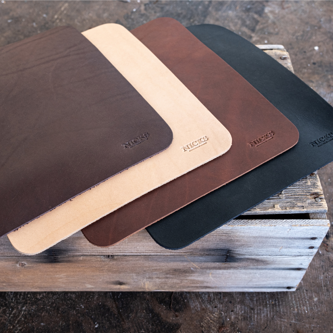 Four leather mouse pads in dark brown, light tan, medium brown, and black, arranged in a fan shape on a rustic wooden crate. The brand name 'NICKS' is embossed on the bottom right corner of each pad.