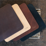 Four leather-like rectangular pads or sleeves in dark brown, tan, reddish brown, and black, arranged overlapping on a rustic wooden surface. Each piece has a small embossed logo near the lower right corner.