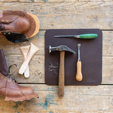 A dark brown leather shoe repair mat laid on a rustic wooden table, surrounded by leatherworking tools including a claw hammer, a green-handled knife, a chisel, nails, and brown leather pieces.