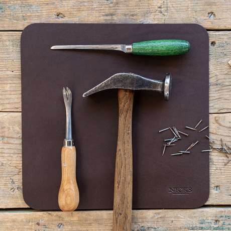 A dark brown leather work mat on a rustic wooden table, featuring a claw hammer, a chisel with a green handle, a nail file, and scattered nails. The brand name 'NICKS' is embossed in the bottom right corner of the mat.