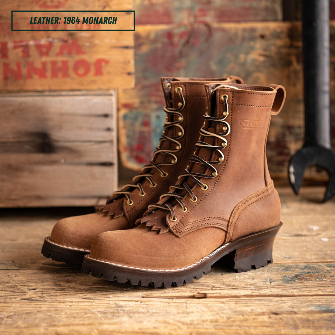 A pair of brown leather lace-up work boots with fringe detailing and metal eyelets, displayed on a rustic wooden surface with vintage crates in the background. A text overlay in the top-left corner reads 'LEATHER: 1964 MONARCH'.