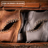 Two work boots displayed inside an open leather trunk: a brown boot on the left and an ash gray boot on the right. Text at the top reads 'LEATHER: 1964 MONARCH & 1964 ASH GRAY'.