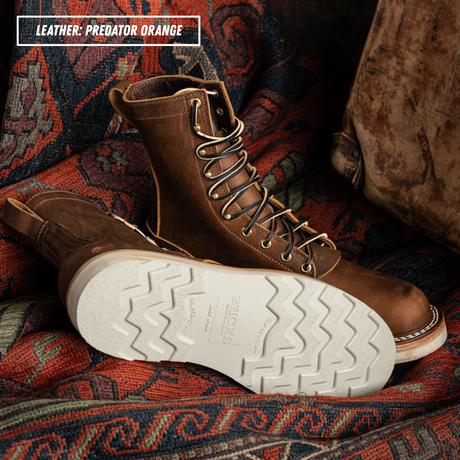 A pair of rugged lace-up work boots in a rich brown leather, with one boot standing upright and the other lying on its side to show the treaded sole. The boots are placed on a patterned red and blue rug with a brown leather bag in the background.