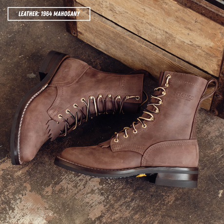A pair of brown suede lace-up boots with gold-tone eyelets and pull tabs, displayed on a rustic concrete floor next to a wooden crate. A text overlay in the top left reads 'LEATHER: 1964 MAGOHANY'.