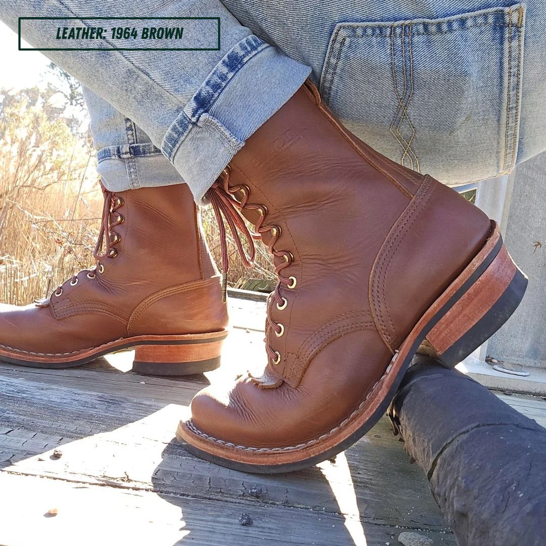 A pair of brown leather lace-up boots worn with cuffed blue jeans, photographed outdoors on a wooden surface. A text overlay in the top left reads 'LEATHER: 1964 BROWN'.