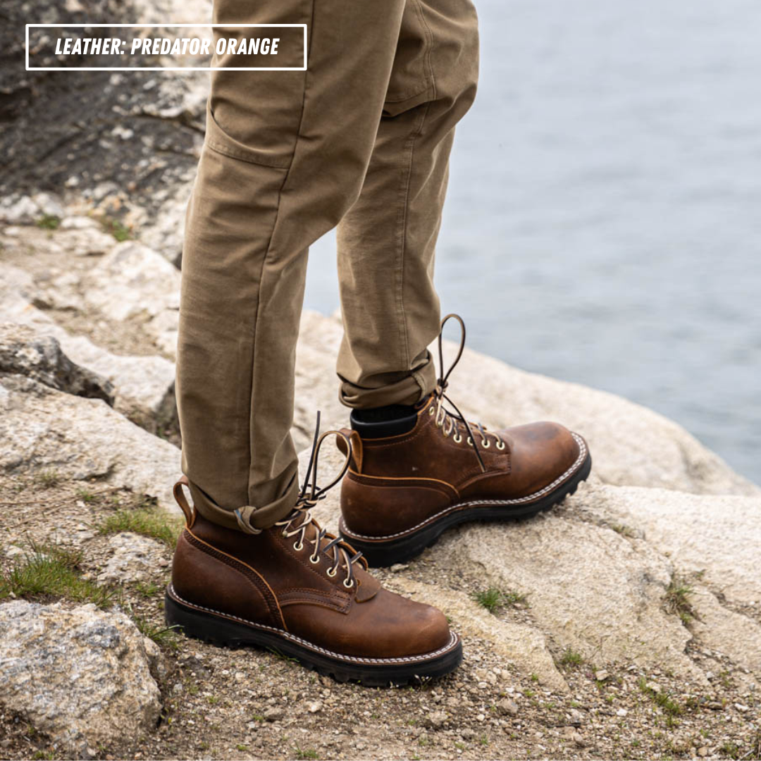 A person wearing brown leather lace-up boots with tan pants, standing on a rocky shoreline with water in the background. A text overlay in the top left reads 'LEATHER - Predator Orange'.
