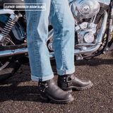 A person wearing cuffed light blue jeans and black leather motorcycle boots stands beside a chrome motorcycle on asphalt. A text overlay in the top left reads 'LEATHER: SHRUNKEN BISON BLACK'.