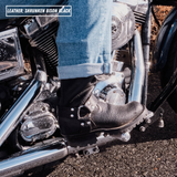 A close-up lifestyle shot of a person's foot wearing a black leather motorcycle boot with silver studs, resting on the footpeg of a chrome motorcycle. The person is wearing blue jeans, and the background shows asphalt and the motorcycle engine.