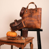 Cypress moc tote sitting on black stool with matching belt hanging off corner of bag in front of warm white background. Various leather goods including an Every Day Carry case, catch all, and drink sleeve sit on wooden table just below.