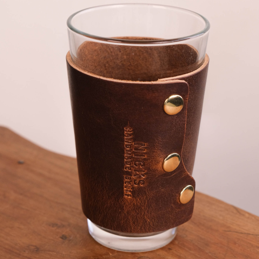 A brown leather pint glass sleeve with brass snap buttons and vertical embossed branding, shown on a clear glass tumbler resting on a wooden surface against a light neutral background.