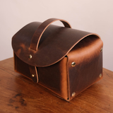 A rustic brown leather storage case with a top handle and brass rivets, resting on a wooden table against a plain light background.
