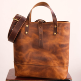 A brown leather tote bag sitting on brown wooden stool in front of off white background. Matching belt hanging off tote.