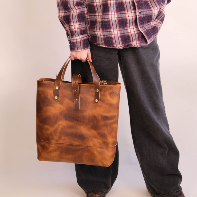 A person wearing a red and navy plaid flannel shirt and dark gray jeans holds a large, distressed brown leather tote bag with brown leather handles and brass hardware against a plain off-white background.