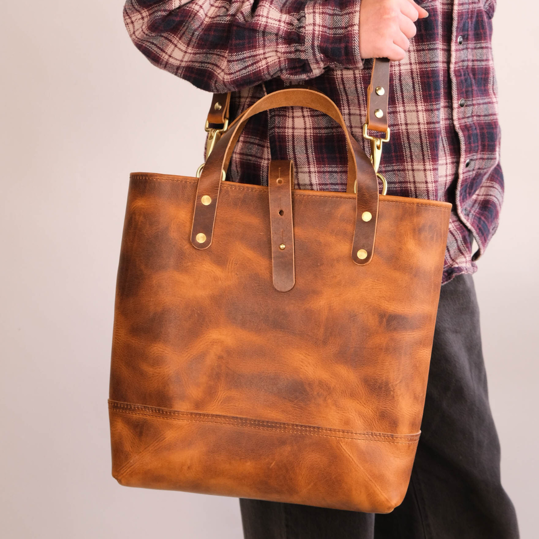 A person wearing a red and blue plaid shirt carries a large brown leather tote bag with gold-tone hardware against a plain light gray background.