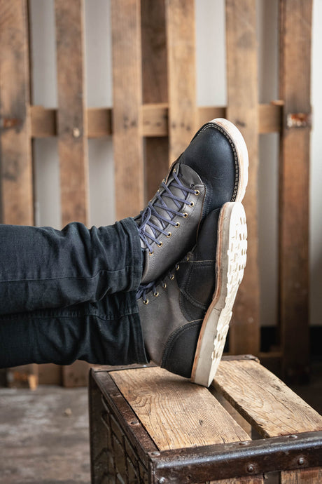 Person wearing black boots with white soles on a wooden crate against a wooden wall.