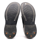 A pair of black leather boots shown from the sole side, featuring textured tread patterns, white contrast stitching, and orange lug nuts on a white background.