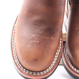 Close-up of a brown leather boot showing the vamp with visible scuffs and scratches on the leather surface, white contrast stitching around the edge, and part of the sole.