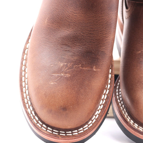 Close-up of a brown leather boot showing the vamp with visible scuffs and scratches on the leather surface, white contrast stitching around the edge, and part of the sole.