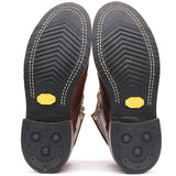 A pair of brown leather boots shown from the sole view, displaying the treaded black rubber soles with white stitching and yellow Vibram logos.