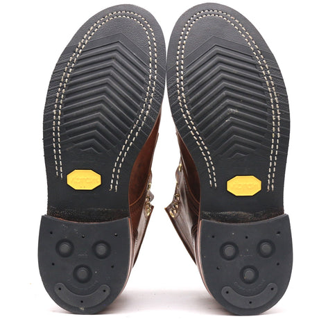 A pair of brown leather boots shown from the sole view, displaying the treaded black rubber soles with white stitching and yellow Vibram logos.
