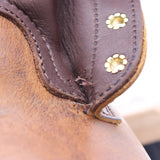 Close-up of a brown leather item showing stitched leather panels, a small tear or frayed edge, and two gold-tone flower-shaped rivets or studs.
