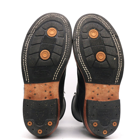 The soles of a pair of black boots, showing the tread pattern, white contrast stitching, and brown leather midsoles with metal cleats.