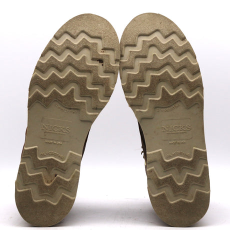 A pair of boots shown from the bottom, displaying their textured zig-zag treaded soles. The soles are a weathered tan-brown color with the brand name 'NICKS' clearly embossed in the center.