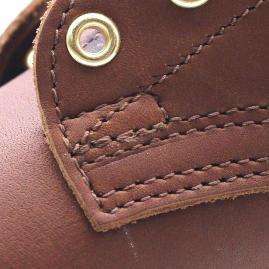 Close-up detail of a brown leather item showing stitched panels, contrast stitching, and a brass snap button.
