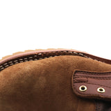 Close-up of a brown suede boot or shoe showing the suede texture, contrast stitching, and a brown leather strap with brass eyelets against a white background.