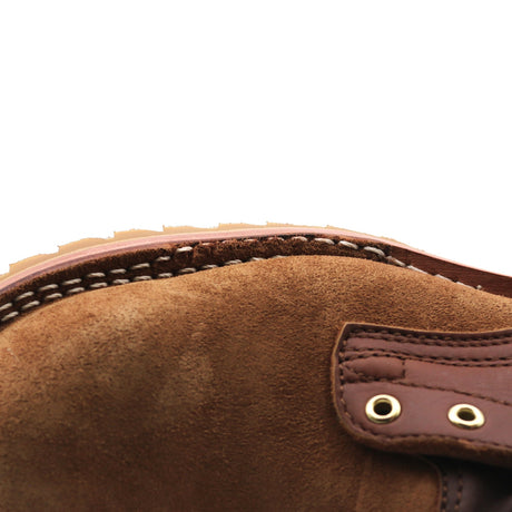 Close-up of a brown suede boot or shoe showing the suede texture, contrast stitching, and a brown leather strap with brass eyelets against a white background.