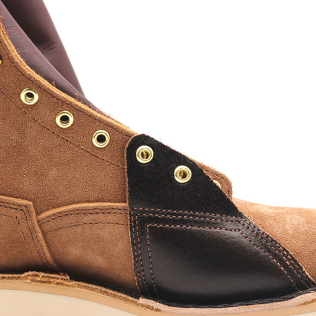 Close-up of a brown suede and black leather ankle boot with brass eyelets on a white background.
