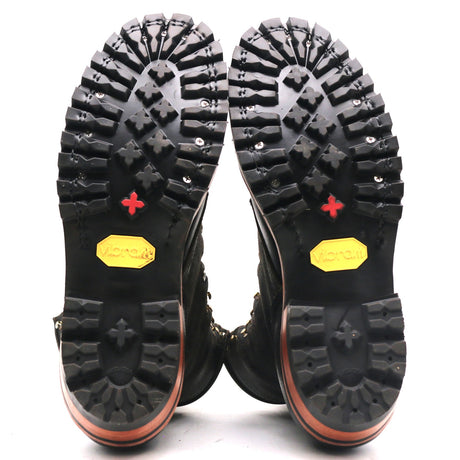A pair of black boots shown from the sole view, featuring deep treaded rubber soles with a red cross symbol and a yellow Vibram logo, set against a plain white background.