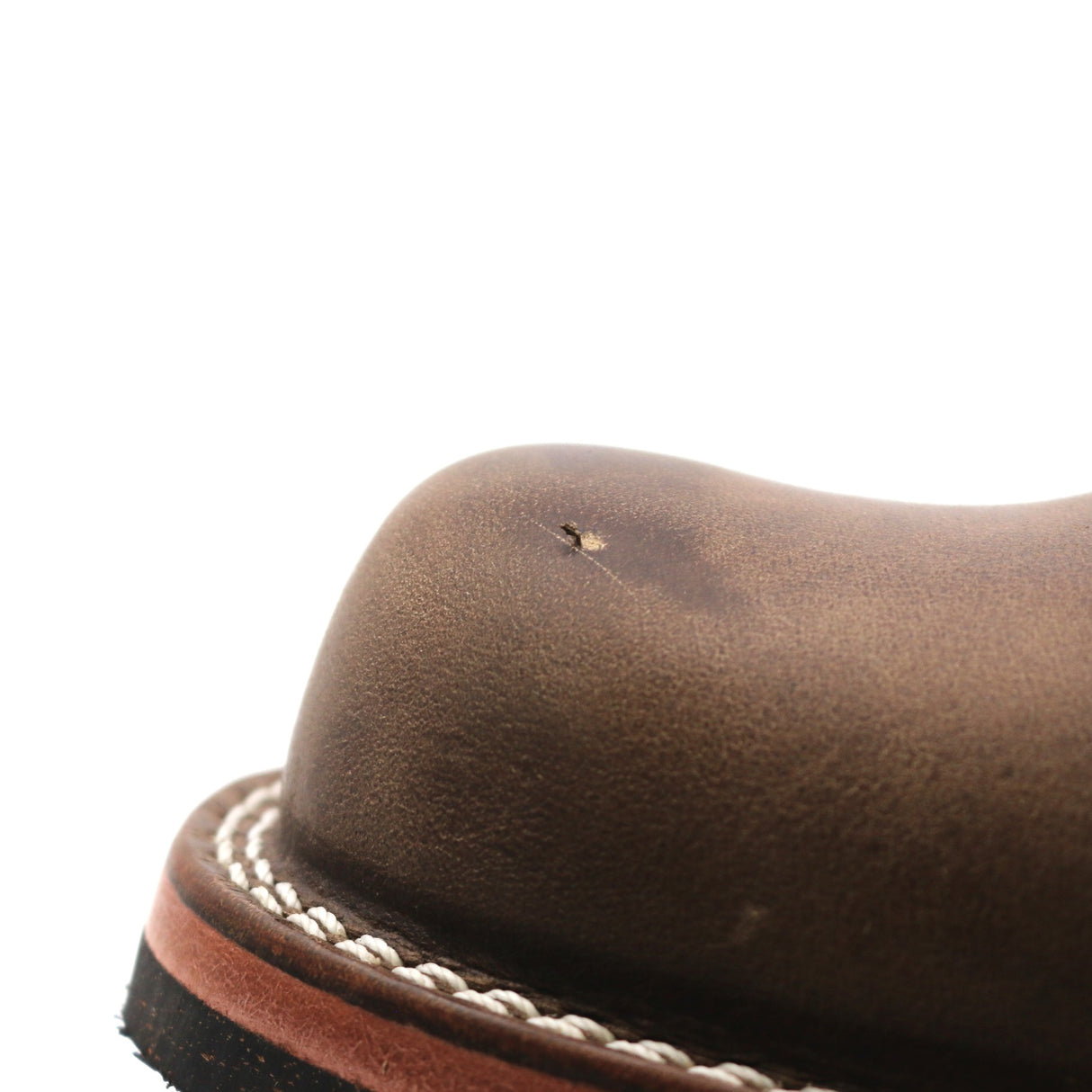 Close-up of a brown leather boot toe showing a small surface defect and white contrast stitching along the edge of a dark sole, on a white background.