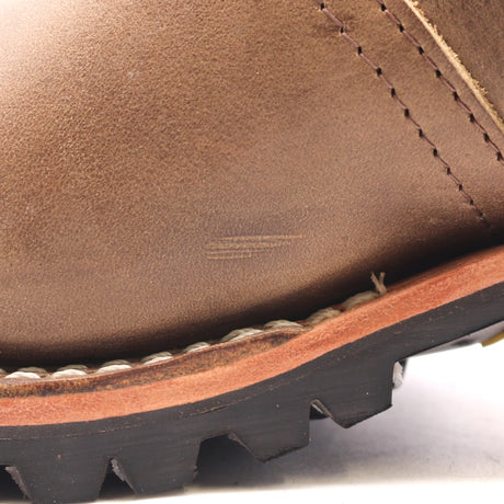 Close-up detail of a brown leather boot showing the side panel, stitching, a tan midsole edge, and a black lug sole.