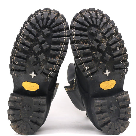 A pair of black boots shown from the sole view, displaying deep lugs, white contrast stitching, and yellow Vibram soles on a white background.