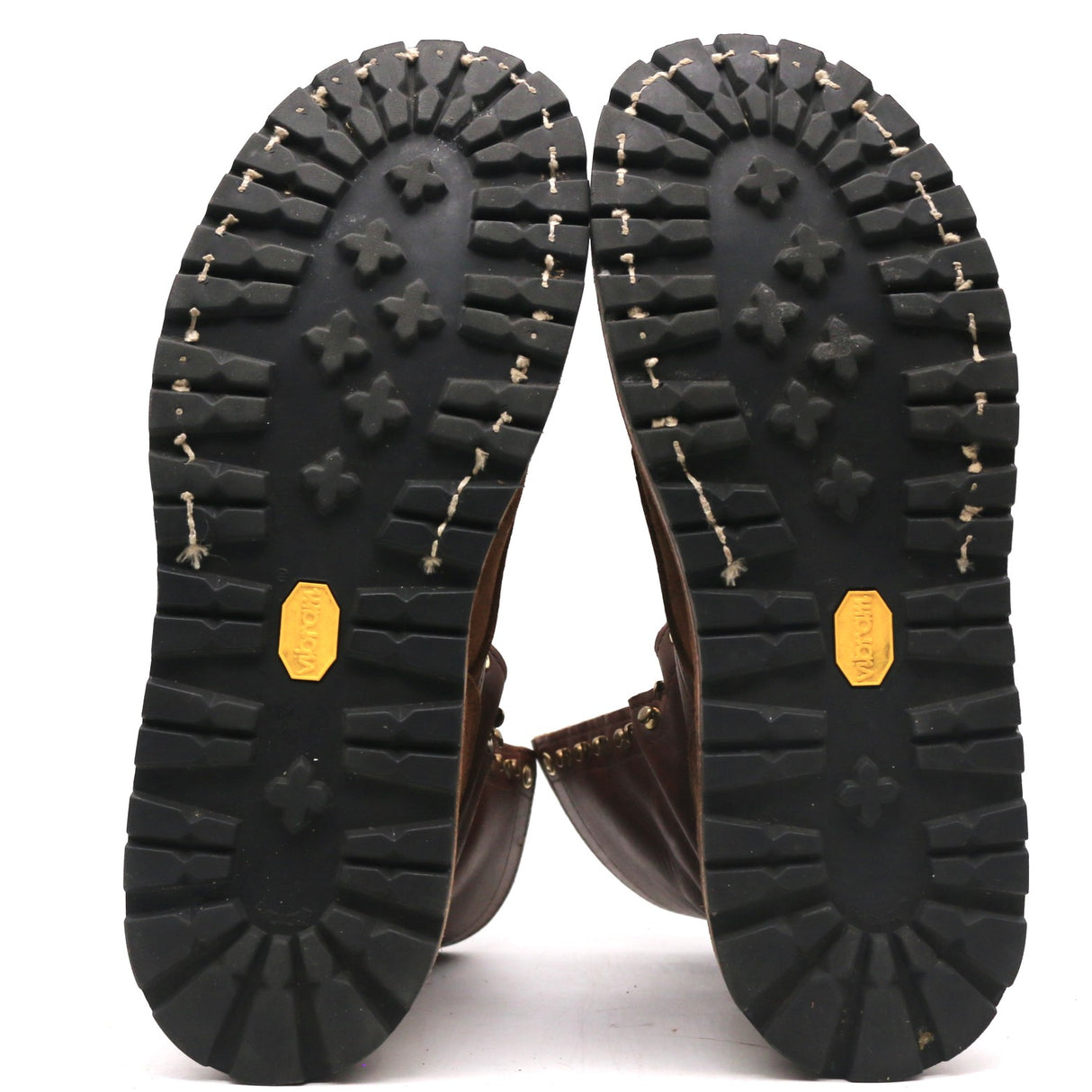 The soles of a pair of brown leather boots, showing deep black tread patterns, whipstitching, and yellow Vibram logos on a white background.