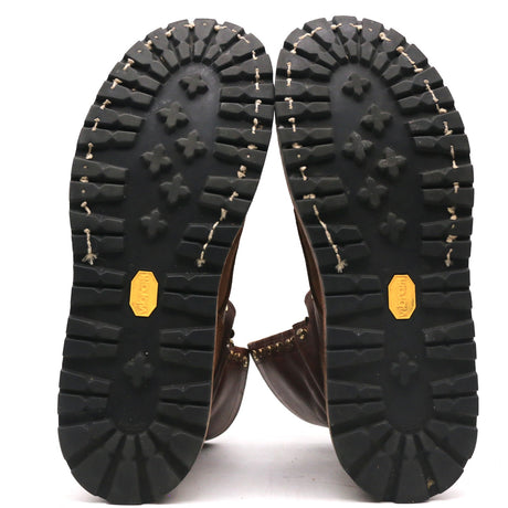 The soles of a pair of brown leather boots, showing deep black tread patterns, whipstitching, and yellow Vibram logos on a white background.