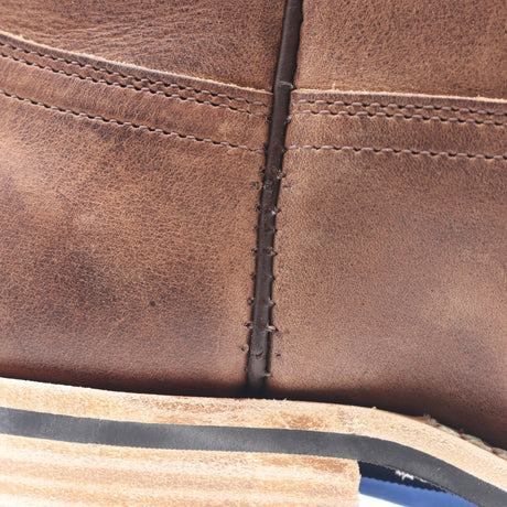 Close-up detail of a brown leather boot showing the texture of the leather, decorative stitching, and a portion of the layered wooden heel.