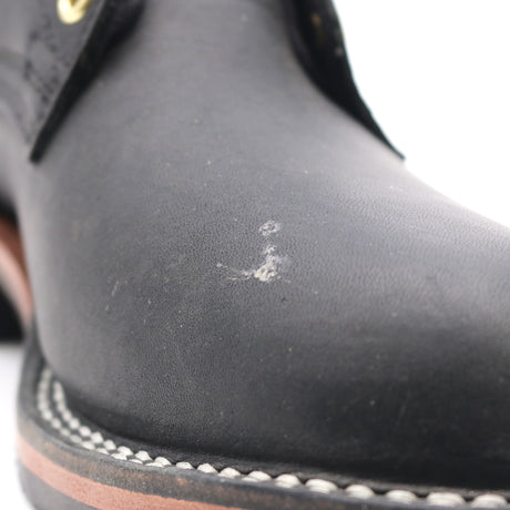 Close-up of a black leather boot toe showing a small white scratch or scuff on the vamp, with white stitching and a brown outsole edge visible against a white background.