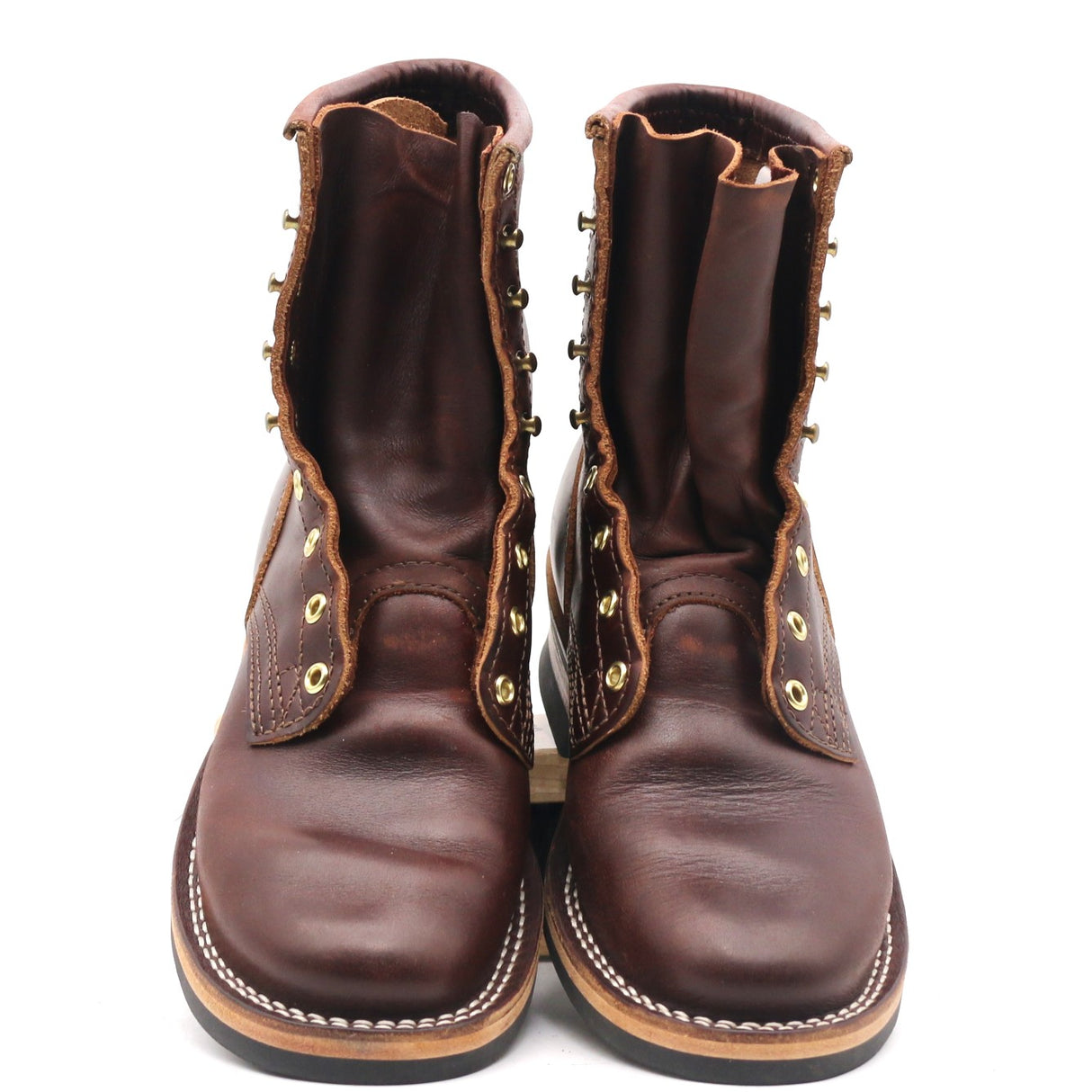A pair of dark brown leather lace-up boots with brass grommets and white contrast stitching, shown from the front against a white background.