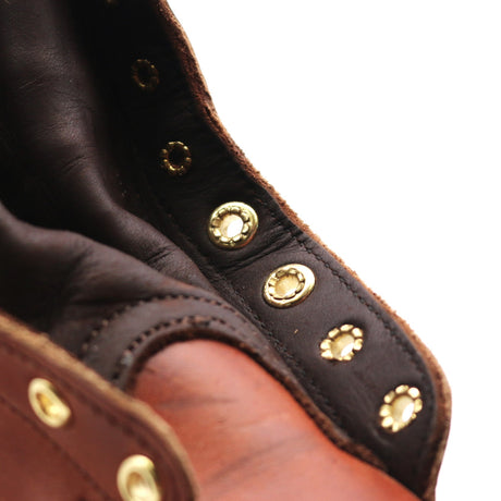 Close-up of a leather boot or shoe showing dark brown leather trim, a tan leather section, and brass-tone metal eyelets against a white background.