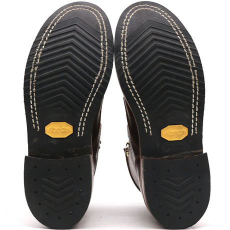 The soles of a pair of dark brown leather boots, showing the black treaded rubber outsoles with white contrast stitching and yellow Vibram logos.